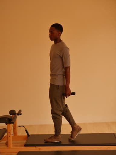 이 이미지는 편안한 운동복과 슬리퍼를 착용한 30대 중반의 아프리카계 남성이 따뜻한 필라테스 스튜디오에서 필라테스 매트를 배경으로 부드럽게 춤추는 모습을 담고 있으며, 편안함과 차분함을 표현하고 있습니다.