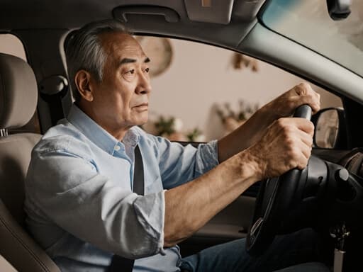 초반 50대 아시아계 남성이 자연스러운 외모로 캐주얼한 셔츠와 청바지를 입고 거실에 앉아 핸들을 불안하게 잡고 있는 반신 샷. 아늑한 배경이 그의 불안한 표정을 보완하여 친근하고 친밀한 분위기를 만들어냅니다.