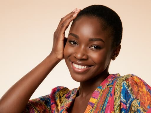 매력적인 20대 초반의 아프리카계 여성의 반신 사진으로, 화려한 패턴의 블라우스를 입고 자연스러운 미소를 지으며 자신감 넘치는 모습을 보여줍니다. 그녀는 스튜디오에서 부드러운 조명이 비추는 가운데, 한 손을 머리에 얹고 화장하는 모습으로 따뜻하고 매력적인 분위기를 연출합니다.