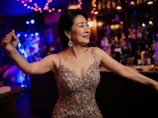 이미지는 40대의 우아한 동양인 여성이 화려한 생일 파티 드레스를 입고 스타일리시한 귀걸이를 착용한 채로 아늑한 바에서 다채로운 조명이 비추는 가운데 우아하게 춤추고 있는 모습을 담고 있으며, 흥분과 매력을 동시에 드러내고 있습니다.