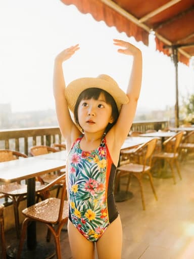 컬러풀한 수영복과 모자를 착용한 7세 아시아계 소녀가 햇살 가득한 카페 테라스에서 두 팔을 위로 쭉 뻗으며 고개를 갸우뚱하고 배고픈 표정을 짓고 있는 모습입니다.
