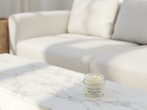 손크림이 잘 닦인 흰색 대리석 표면에 놓여 있으며, 부드러운 자연광이 비추고 세밀한 질감이 드러나며, 미세한 결함이 있고 현대적인 흰색 라운지의 미니멀한 장식이 배경을 이루며 부드러운 그림자가 드리워진 따뜻하고 아늑한 분위기이다.