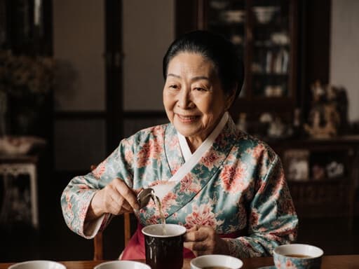 전통적인 한복을 입은 80대 혼혈 여성의 따뜻하고 아늑한 반신 촬영으로, 전통적인 거실의 아늑한 테이블에 앉아 차를 따르며 편안한 미소를 짓고 있습니다. 배경은 문화적인 가구와 소품들로 구성되어 있어 장면의 초대하는 분위기를 강화합니다.
