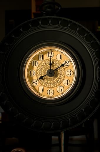 세련된 검은색과 금색 벽시계가 아름답게 디자인된 미니멀리스트 거실에 prominently 걸려 있습니다. 우아한 시계는 복잡한 시계 바늘과 크고 읽기 쉬운 숫자를 포함하고 있습니다. 부드러운 자연광이 방을 가득 채우며 평화로운 분위기를 강조합니다. 부드러운 안락의자와 흐릿한 책장이 배경에 있어 아늑하고 세련된 분위기를 더합니다.