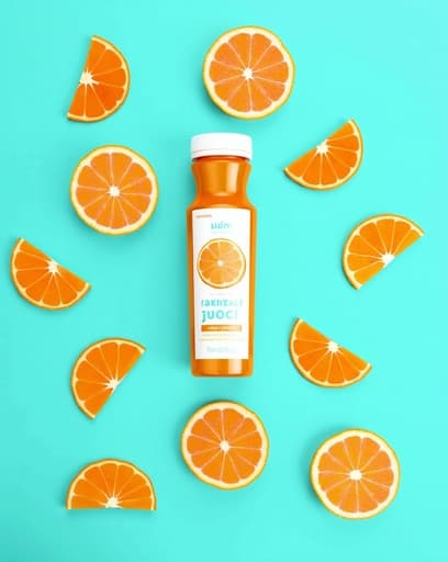 갓 짜낸 오렌지 주스 한 잔이 나무 테이블 위에 놓여 있고, 생생한 오렌지 조각으로 장식되어 있습니다. 배경은 햇빛이 창문을 통해 들어오는 밝은 주방으로, 신선하고 매력적인 분위기를 자아냅니다.