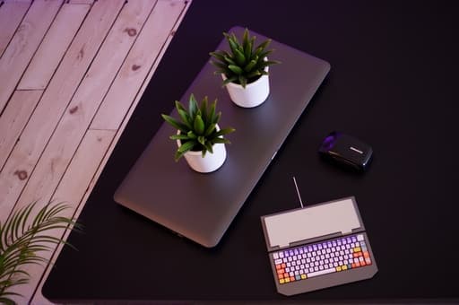 현대적인 홈 오피스의 세련된 책상 위에 커스터마이즈 가능한 RGB 조명이 있는 인체공학적 컴퓨터 키보드의 고품질 이미지가 강조되어 있습니다. 배경에는 노트북과 몇 개의 녹색 실내 식물이 있어 생산적이고 현대적인 분위기를 자아냅니다.