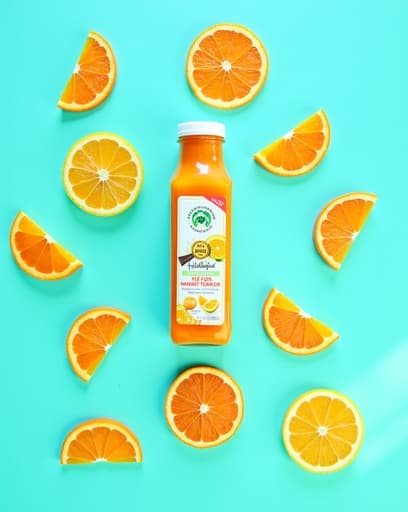 신선하게 squeezed된 오렌지 주스가 나무 테이블 위에 놓여 있으며, 신선한 오렌지 조각으로 장식되어 있습니다. 주방은 밝고 통풍이 잘 되며, 햇빛이 창문을 통해 비추어 주스를 밝은 색으로 비추고 있습니다. 흩어져 있는 오렌지와 도마가 장면에 따뜻한 가정적인 느낌을 더합니다.