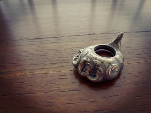 잘 다듬어진 금속 물체가 광택 있는 나무 테이블 위에 놓여 있는 클로즈업 샷입니다. 이 물체는 반사되는 마감 처리가 되어 있어, 그 품질을 강조하는 복잡한 디자인을 보여줍니다. 부드럽게 초점이 흐려진 배경은 따뜻하고 초대하는 방을 연상시키며, 금속 조각의 우아함을 더해줍니다.