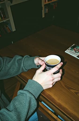 한 사람이 아늑한 카페에서 아름답게 디자인된 세라믹 커피 컵을 들고 있습니다. 부드러운 조명이 따뜻한 분위기를 만들어주며, 배경에는 나무 테이블과 책으로 가득 찬 책장이 있어 장면의 초대하는 느낌을 더해줍니다.