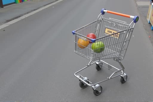 반짝이는 금속 쇼핑 카트가 바쁜 야외 쇼핑 지역의 바닥에 놓여 있으며, 주변에는 다채로운 상점들과 쇼핑을 즐기는 사람들로 가득한 화창한 날의 분위기가 느껴진다. 카트는 넓은 바구니와 네 개의 튼튼한 바퀴를 가지고 있어 식료품이나 다른 구매물로 가득 채워질 준비가 되어 있다.