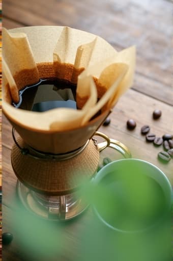 친환경 커피 필터가 나무 테이블 위에 놓여 있고, 주변에는 흩어져 있는 커피 원두와 방금 내린 따뜻한 커피 잔이 있으며, 부드러운 자연광이 장면을 밝혀주는 모습의 클로즈업.