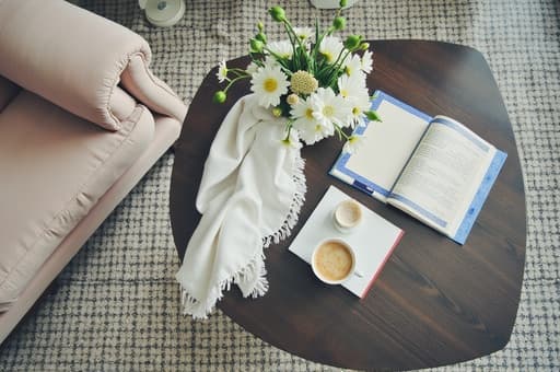 스타일리시한 커피 컵과 열린 책이 놓인 아름답게 정돈된 커피 테이블. 광택이 나는 나무 테이블은 아늑한 거실의 따뜻하고 부드러운 조명을 반사하며, 푹신한 소파와 자연광이 들어오는 창문이 초대하는 분위기를 만들어낸다.