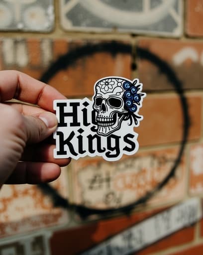 한 사람이 생동감 넘치는 도시 배경의 벽화가 그려진 벽 앞에서 스타일리시한 해골 스티커를 들고 있다. 스티커의 세부 사항과 디자인에 초점이 맞춰져 있으며, 배경은 강렬한 분위기를 더한다.