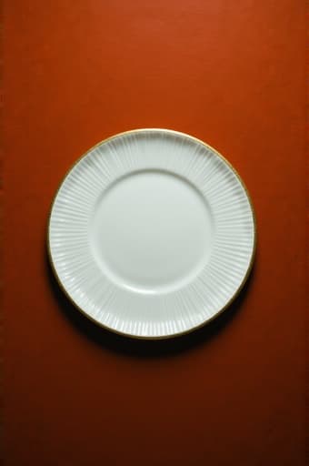 반짝이는 금 테두리가 있는 아름답게 배열된 흰색 접시가 깊은 빨간색 표면 위에 두드러지게 놓여 있어 우아함과 고급스러움을 발산합니다. 깨끗한 접시와 풍부한 배경 간의 대비가 놀라운 시각적 매력을 만들어 내며, 고급 다이닝 설정이나 장식 디스플레이에 적합합니다.