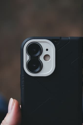 한 사람이 세련된 검은색 휴대전화 케이스를 한 손으로 들고 있는 클로즈업 이미지로, 흐릿한 현대 도시 배경이 보입니다. 이 휴대전화 케이스는 내구성이 뛰어난 디자인과 정확한 컷아웃으로 기능성과 스타일을 강조하고 있습니다. 그 사람의 손은 우아하게 위치해 있어 자신감과 트렌디함을 암시합니다.