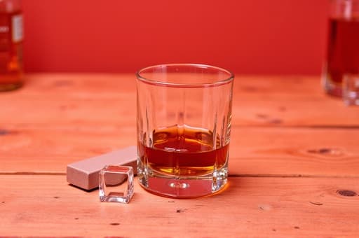 잘 만들어진 크리스탈 위스키 잔에 깊고 풍부한 호박색 위스키가 담겨 있으며, 거친 나무 바닥 위에 놓여 있습니다. 얼음 조각이 시원함을 더하고, 위스키 스톤이 잔 옆에 있습니다. 부드러운 조명의 따뜻한 빛이 아늑한 분위기를 만들어내고, 배경에 흐릿하게 보이는 위스키 병들이 장면의 세련된 느낌을 더욱 강조합니다.