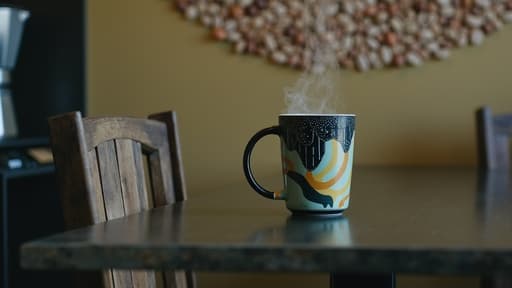아늑한 카페의 러스틱한 나무 테이블 위에 아름답게 디자인된 세라믹 커피 컵이 놓여 있습니다. 컵은 현대적인 디자인을 보여주며 생생한 색상을 자랑합니다. 배경에는 부드럽게 흐려진 커피 원두와 김이 나는 커피 포트가 있어 따뜻하고 초대하는 분위기를 연출합니다.