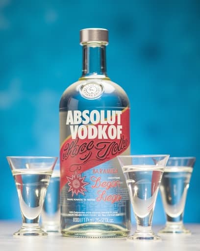 세련된 바 카운터 위에 아름답게 배열된 Absolut 보드카 병이 우아한 샷 글라스와 함께 자리잡고 있습니다. 부드러운 조명이 따뜻하고 초대하는 분위기를 연출하며, 고급스러운 모임에 적합합니다. 배경은 흐릿한 바 풍경을 보여주어 세팅의 고급스러운 느낌을 더합니다.