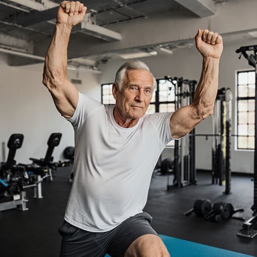 넓은 헬스장에 있는 건강하고 활기찬 50대 백인 남성이 스트레칭을 하며 자신감 있는 표정을 짓고 있는 반신 샷. 편안한 스포츠웨어를 착용하며, 활동적인 라이프스타일을 보여줍니다.