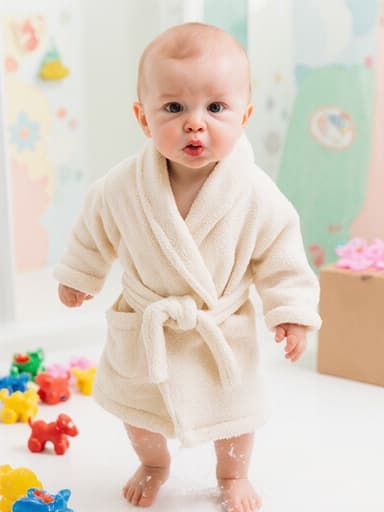 부드러운 면 소재의 목욕 가운을 입은 1세의 유럽계 남성 아기가 목욕을 마친 후 물기를 털어내며 장난스럽게 걷고 있는 전신 사진입니다. 그는 짜증난 표정을 지으며 입술을 쭉 내밀고 있습니다. 배경은 밝고 아기자기한 욕실로, 물놀이 장난감이 흩어져 있어 장난스럽고 사랑스러운 순간을 담고 있습니다.