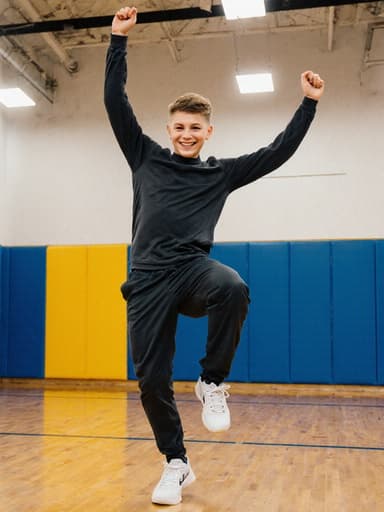 10대 초반의 백인 남성이 편안한 운동복을 입고 밝은 체육관에서 즐겁게 춤추며 팔을 높이 들고 환하게 웃고 있는 모습으로, 재미와 활력을 상징하고 있습니다.