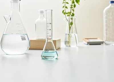 투명한 유리 비커가 선명한 파란색 액체로 가득 차 있으며, 세련된 실험실 작업대 위에 놓여 있습니다. 비커는 정확한 측정 눈금이 특징이며, 배경에는 과학 기구와 몇 개의 녹색 식물이 있어 부드럽고 확산된 조명에 의해 비추어져 실험과 혁신의 분위기를 만들어냅니다.