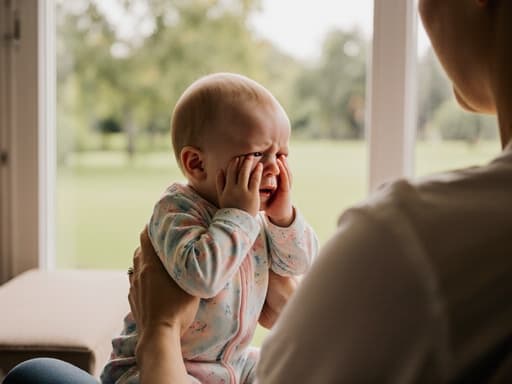 이 이미지는 편안한 파스텔 색상의 수면복을 입은 1세 유럽계 여성 아기가 부모의 품에서 앉아, 슬픈 표정을 지으며 손으로 얼굴을 가리고 있는 모습을 담고 있습니다. 부드러운 햇살이 비치는 거실과 창문 너머의 녹색 공원이 부드럽고 감정적인 분위기를 만들어냅니다.
