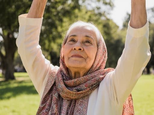 전통적인 의상을 입은 중동계 노인 여성의 반신 사진으로, 화려한 스카프를 두르고 햇살이 비치는 공원에서 스트레칭하며 여유롭고 즐거운 표정을 짓고 있습니다.