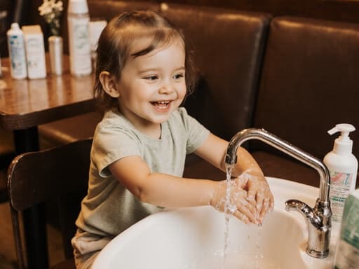 아늑한 카페 테이블에 앉아 있는 귀여운 3세 백인 소녀의 반신 사진입니다. 밝은 색상의 티셔츠와 편안한 바지를 입고 있으며, 행복한 미소를 지으며 손을 씻고 있습니다. 테이블 위에는 손 씻기 용품이 놓여 있으며, 카페의 따뜻하고 초대하는 분위기가 즐거운 상호작용을 강조합니다.