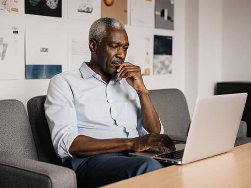 50대의 흑인 남성이 회의실의 현대적인 소파에 편안하게 앉아 있는 모습입니다. 그는 캐주얼한 셔츠를 입고 자연스러운 표정을 지으며 노트북을 작업하고 있습니다. 벽에는 다양한 디자인 샘플이 걸려 있어 창의적이고 편안한 분위기를 전달합니다.