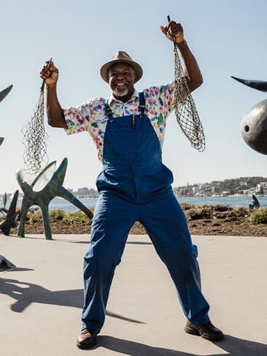 60대 흑인 남성이 어부 스타일의 작업복과 모자를 착용하고, 해양 생태 보호 공원에서 낚시 도구를 들고 춤추며 활기차고 즐거운 표정을 짓고 있는 반신 샷, 바다 관련 조형물로 둘러싸인 모습이 생동감 넘치고 에너지를 전달합니다.
