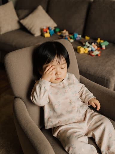 부드러운 색상의 잠옷을 입고 졸린 표정으로 눈을 비비고 있는 1세 한국인 여성 아기의 반신 촬영. 안락의자에 살짝 앞으로 기울여 앉아 있으며, 주변은 장난감이 흩어져 있는 아늑한 거실과 편안한 소파로 둘러싸여 있어 유아의 편안함과 평온함이 느껴지는 완벽한 분위기를 만들어냅니다.