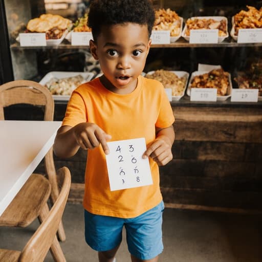 아늑한 카페에서 7세 남성 혼혈 아동이 밝은 색의 티셔츠와 반바지를 입고, 배고픈 표정을 지으며 숫자가 적힌 카드를 들고 식사와 음료가 가득한 테이블을 가리키고 있습니다.