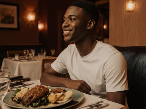 친근한 미소를 지으며 즐거운 감정을 표현하는 10대 후반의 흑인 남성이 캐주얼한 티셔츠와 청바지를 입고 레스토랑 테이블에 앉아 있습니다. 그는 테이블에 놓인 다양한 맛있는 음식을 바라보며 편안하게 앉아 있습니다. 아늑한 레스토랑 내부의 따뜻한 조명과 잘 정돈된 테이블 세팅이 따뜻한 분위기를 연출합니다.