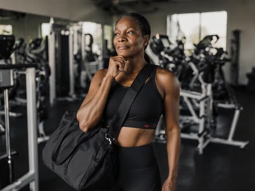 운동복을 입고 운동 가방을 멘 50대 흑인 여성의 반신 사진으로, 넓은 운동 공간과 다양한 운동 기구가 있는 헬스장에서 건강 관리에 대해 깊은 생각에 잠긴 모습이 강조됩니다.