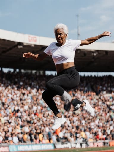 60대 아프리카계 여성의 전신 사진으로, 편안한 운동복을 입고 무관심한 표정으로 점프하는 모습이 담겨 있습니다. 배경에는 관중들이 가득한 활기찬 운동 경기장이 있어 장면에 에너지를 더하고 그녀의 역동적인 움직임을 강조합니다. 전체적인 분위기는 캐주얼한 자신감을 전달하며, 그녀의 자연스러운 활동 참여를 보여줍니다.