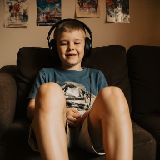 7세 백인 남성이 자연스러운 외모로 편안한 티셔츠와 반바지를 입고 음악을 집중해서 듣고 있는 모습의 반신 촬영. 그의 표정은 배고픈 듯 하며, 아늑한 거실에서 다리를 뻗고 앉아있고, 벽에 걸린 포스터와 소파가 있는 따뜻하고 초대하는 분위기를 연출하고 있습니다.