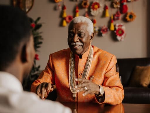 이 이미지는 화려한 전통 축제 의상을 입고, 회의실 테이블에 앉아 주변 사람들과 즐겁게 소통하는 친근한 표정을 지닌 80대 아프리카계 남성을 담고 있습니다. 축제로 장식된 거실 배경에서 그의 자연스러운 제스처는 따뜻한 공동체 의식을 전달합니다.