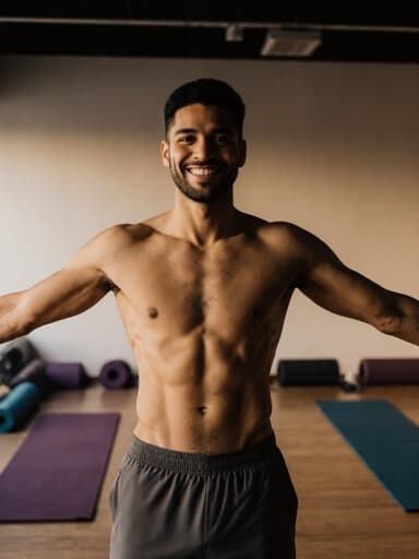 30대 라틴계 남성이 편안한 요가복을 입고 매력적인 미소를 지으며, 아늑한 요가 센터에서 자연스럽게 팔을 옆으로 뻗고 서 있는 반신 촬영. 부드러운 조명 아래에서 차분한 분위기를 더하고 있습니다.