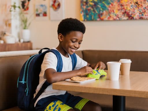 아늑한 카페에서 호기심 가득한 표정으로 가방을 들여다보는 사랑스러운 7살 아프리카계 남성의 반신 사진. 편안한 티셔츠와 반바지, 밝은 색상의 운동화를 신고 있으며, 배경은 예술작품과 음료, 간식으로 꾸며져 있습니다.