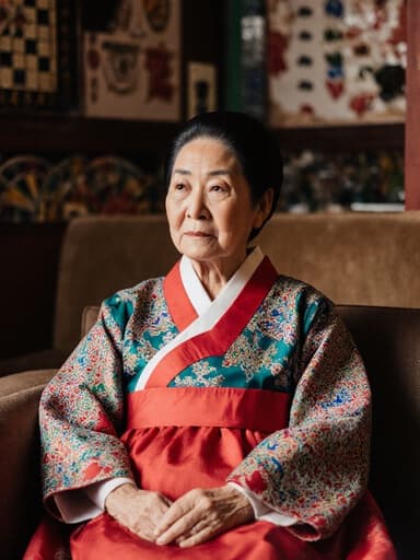 70대 한국인 여성이 아늑한 카페의 소파에 편안하게 앉아 있는 반신 사진입니다. 그녀는 화려하고 정갈한 전통 한복을 입고 있으며, 무관심한 표정으로 주변을 바라보며 전통 놀이를 주제로 한 장식이 있는 카페의 분위기를 즐기고 있습니다. 전체적으로 차분하고 초대하는 느낌의 이미지입니다.