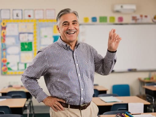 50대의 행복한 라틴계 남성 교사가 체크 무늬 셔츠와 면바지를 입고 밝은 교실에서 한 손은 허리에, 다른 손은 학생들과 함께 춤추는 듯한 자세로 서 있으며, 정돈된 책상과 교육 자료로 둘러싸여 교육에 대한 열정과 따뜻함을 드러내고 있습니다.