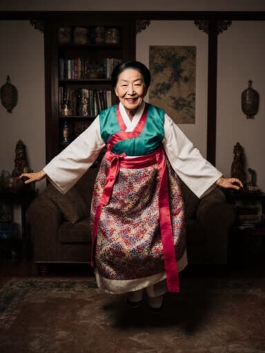 전통 한복을 입고 있는 70대 동양인 여성의 전신 사진으로, 고마워하는 표정을 지으며 아늑한 거실에서 점프하고 있습니다. 따뜻한 조명이 그녀의 기쁜 표정과 의상의 우아함을 강조합니다.