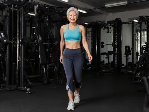 60대 아시아계 여성이 편안한 운동복과 운동화를 입고 세련된 모습으로 자신감 넘치게 걷고 있는 반신 사진입니다. 다양한 운동 기구가 배치된 스포츠 훈련장 안에서 자연스럽게 움직이며 스타일과 자신감을 발산하고 있습니다.