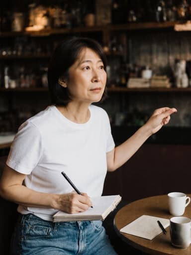 40대 동양인 여성이 편안한 청바지와 티셔츠를 입고 노트를 들고 점프하는 모습을 담은 전신 사진. 그녀는 지루한 표정으로 아늑한 카페를 바라보며 생각에 잠겨 있으며, 테이블 위에는 커피 잔과 책이 있어 편안하면서도 역동적인 분위기를 연출하고 있습니다.