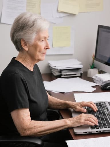 60대 백인 여성의 반신 사진이 정돈된 사무실 환경의 책상 앞에 앉아 있는 모습입니다. 그녀는 편안한 티셔츠와 바지를 입고 있으며, 키보드에 손가락을 얹고 집중한 표정을 짓고 있습니다. 책상 위에는 문서와 노트북이 깔끔하게 정리되어 있어 그녀의 일에 대한 몰입을 강조합니다. 전체적인 분위기는 전문적이고 차분하여 그녀의 집중과 헌신을 부각시킵니다.