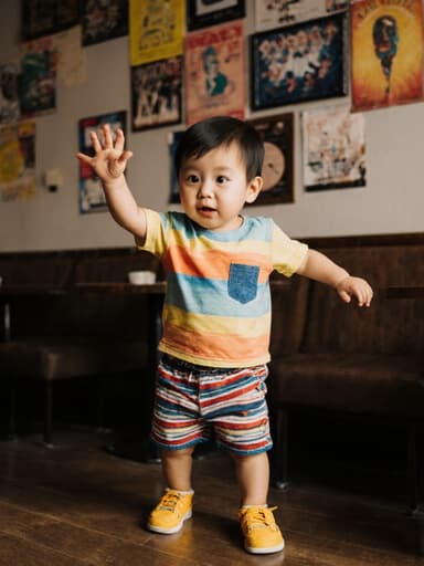 아늑한 카페에서 호기심 어린 표정으로 춤추는 1살 동양인 남아의 전신 촬영, 귀여운 티셔츠와 반바지, 밝은 색상의 신발을 착용하고, 벽에 음악 관련 포스터가 장식되어 있는 모습.