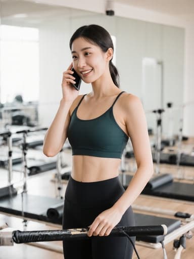 이 이미지는 20대 한국인 여성이 편안한 필라테스 복장을 입고 전화를 귀에 대고 있는 모습으로, 당황한 표정과 부드러운 미소를 지으며 필라테스 기구가 배치된 운동 스튜디오에서 촬영되었습니다.