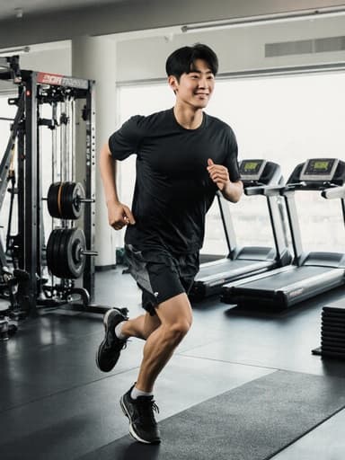 10대 초반의 건강한 외모를 가진 한국인 남성이 헬스장에서 운동복과 트레이닝화를 입고 뛰고 있습니다. 편안하고 자신감 있는 표정이 그의 활동적인 모습과 어우러져 다양한 운동 기구들 사이에서 생동감 넘치는 분위기를 만들어냅니다.