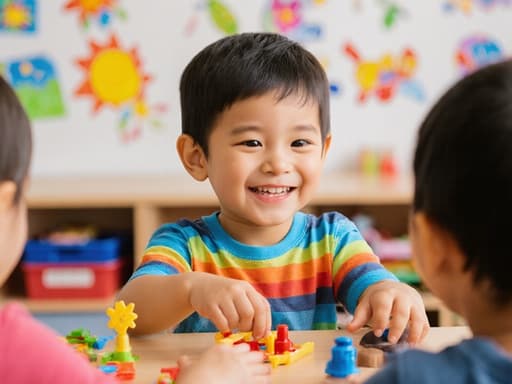 밝은 색의 티셔츠와 반바지를 입은 귀여운 3세 혼혈 아동이 테이블에 앉아 친구들과 함께 장난감을 가지고 놀며 순수한 미소를 지으며 행복해 보이는 모습이 담긴 반신 사진. 아늑한 유치원 교실 배경에는 다양한 학습 자료와 화려한 장식이 있습니다.